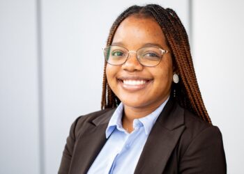 Professional headshot of a smiling Black woman with braided hair, wearing glasses, a light blue shirt and black blazer in an office setting.
