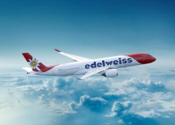 White and red Edelweiss airliner in flight above fluffy clouds, blue sky background, logo on the fuselage.