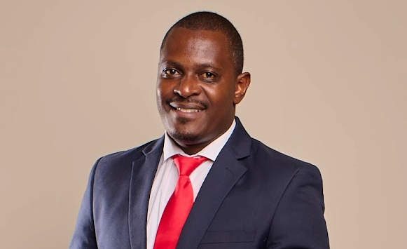 Professional head-and-shoulders portrait of a man in a navy suit and red tie, smiling, against a beige background.