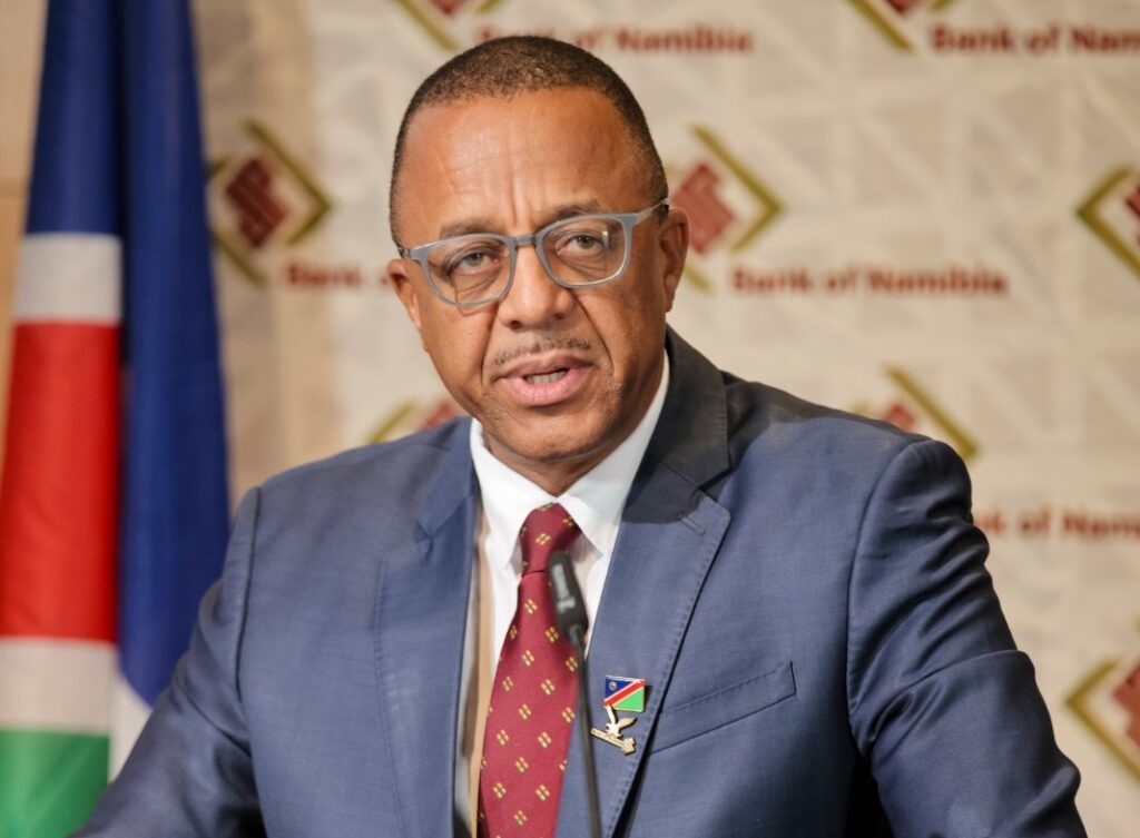 Man in a blue suit and red tie giving a speech at a press conference, Namibian flag in background and a lapel pin visible on his jacket.