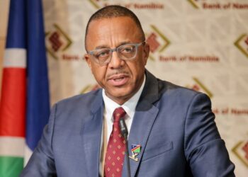 Man in a blue suit and red tie giving a speech at a press conference, Namibian flag in background and a lapel pin visible on his jacket.