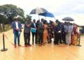 Group of formally dressed people at a ribbon-cutting ceremony outdoors, some holding documents and umbrellas in light rain on a muddy path.