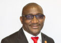Frednard Gideon - The Brief | Namibia's Leading Business & Financial News Professional headshot: smiling man in a black suit, white shirt, and red tie, wearing rectangular glasses against a white background.