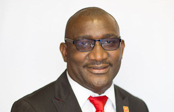 Frednard Gideon - The Brief | Namibia's Leading Business & Financial News Professional headshot: smiling man in a black suit, white shirt, and red tie, wearing rectangular glasses against a white background.
