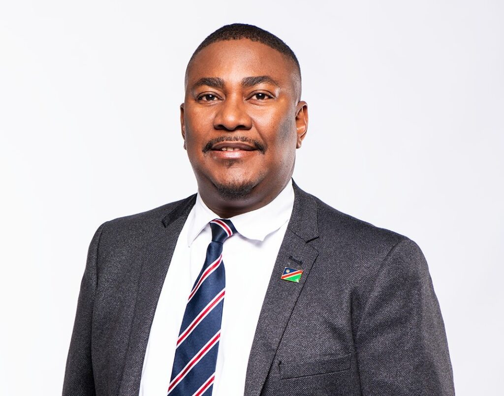 Bolle Hans - The Brief | Namibia's Leading Business & Financial News Head-and-shoulders portrait of a smiling man in a gray suit, white shirt, striped blue-red tie, with a small flag lapel pin on the jacket.