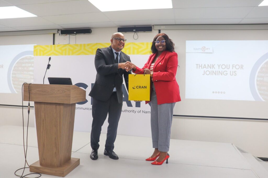 Man in a dark suit and a woman in a red blazer shake hands on a stage while exchanging a yellow CRAN bag; podium left, projector screen thanks attendees in background.