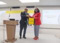 Man in a dark suit and a woman in a red blazer shake hands on a stage while exchanging a yellow CRAN bag; podium left, projector screen thanks attendees in background.