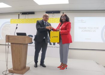Man in a dark suit and a woman in a red blazer shake hands on a stage while exchanging a yellow CRAN bag; podium left, projector screen thanks attendees in background.