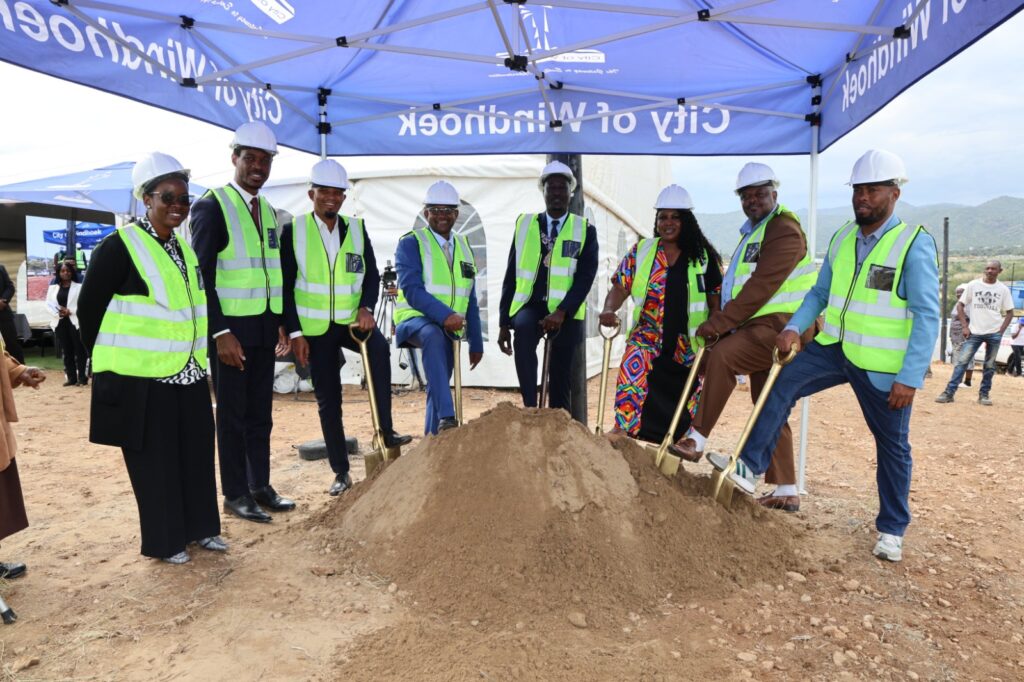 Group of officials in hard hats and neon vests shovel dirt at a ground-breaking ceremony under a blue canopy.
