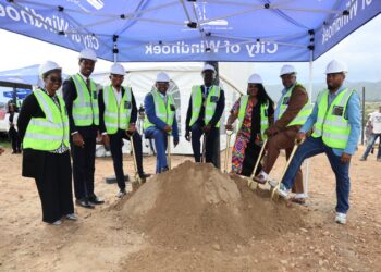 Group of officials in hard hats and neon vests shovel dirt at a ground-breaking ceremony under a blue canopy.