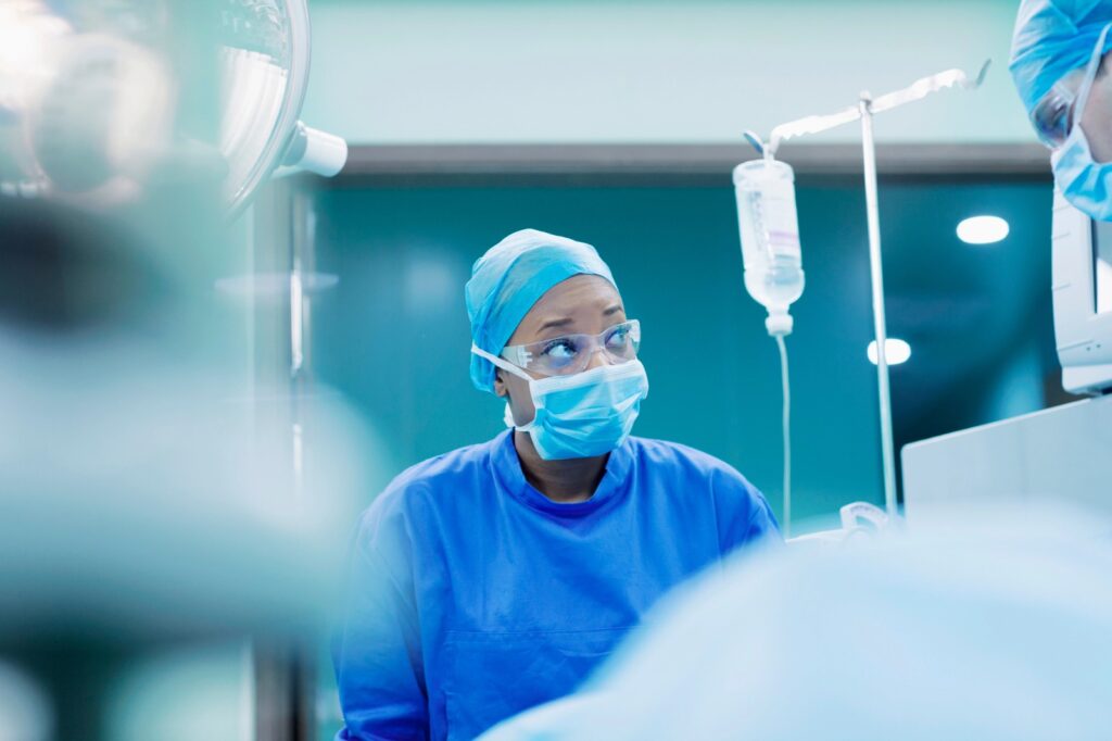 NURSE 1 - The Brief | Namibia's Leading Business & Financial News Surgeon in blue scrubs, mask, and protective goggles in an operating room with an IV drip nearby