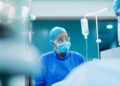 Surgeon in blue scrubs, mask, and protective goggles in an operating room with an IV drip nearby