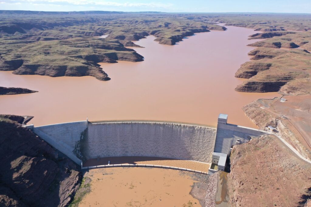 Neckartal Dam - The Brief | Namibia's Leading Business & Financial News Aerial view of a large curved dam holding a muddy brown reservoir in a desert canyon setting now visible from above with rugged hills surrounding the waterway.