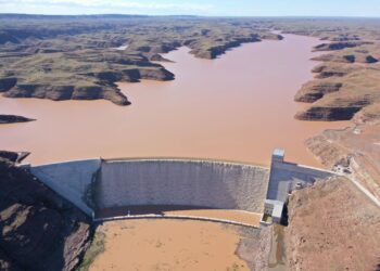 Aerial view of a large curved dam holding a muddy brown reservoir in a desert canyon setting now visible from above with rugged hills surrounding the waterway.