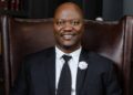 Israel Tjizake - The Brief | Namibia's Leading Business & Financial News Smiling man in a black suit and tie sits in a large brown leather chair with brass studs on the edges.