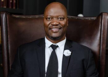 Smiling man in a black suit and tie sits in a large brown leather chair with brass studs on the edges.