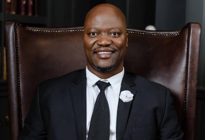 Smiling man in a black suit and tie sits in a large brown leather chair with brass studs on the edges.