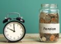 Black retro alarm clock beside a coin-filled jar labeled 'Pension' on a wooden table, illustrating retirement savings.