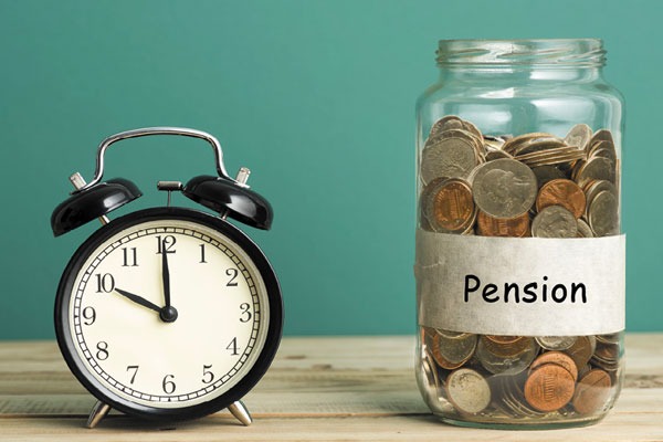 Black retro alarm clock beside a coin-filled jar labeled 'Pension' on a wooden table, illustrating retirement savings.