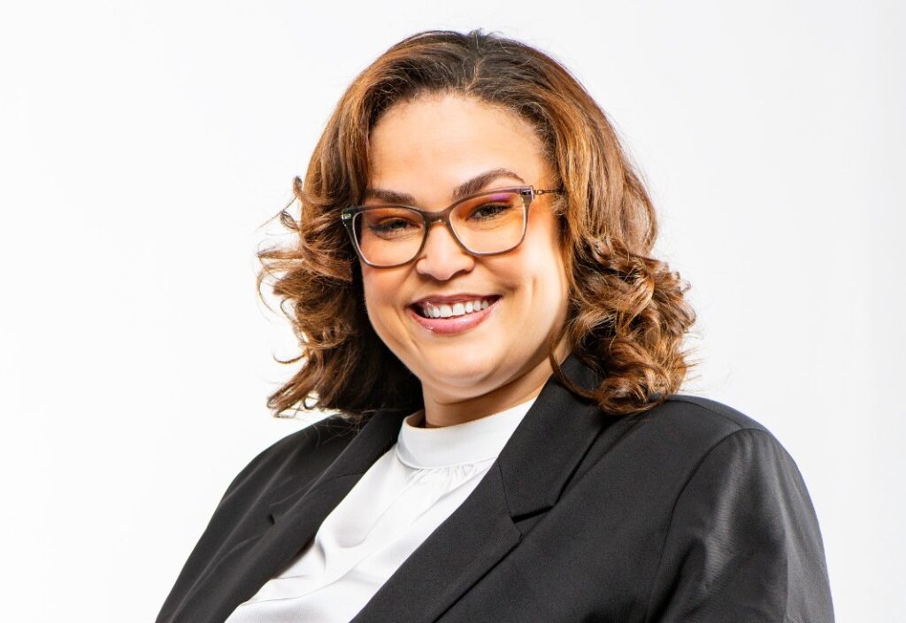 Bronwyn Moody - The Brief | Namibia's Leading Business & Financial News Smiling woman with shoulder-length wavy brown hair, glasses, black blazer, and white blouse against a white background.
