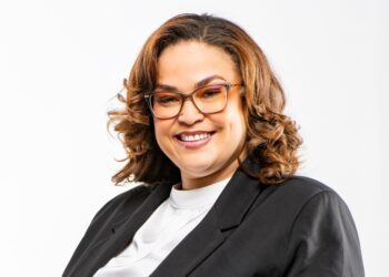 Smiling woman with shoulder-length wavy brown hair, glasses, black blazer, and white blouse against a white background.