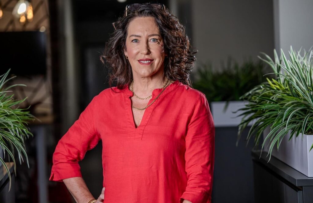 Capricorn Elize Smith 2 1 1 1 - The Brief | Namibia's Leading Business & Financial News Portrait of a smiling woman with curly dark hair wearing a red blouse, standing between potted plants in an office-like setting.
