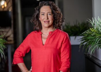Portrait of a smiling woman with curly dark hair wearing a red blouse, standing between potted plants in an office-like setting.