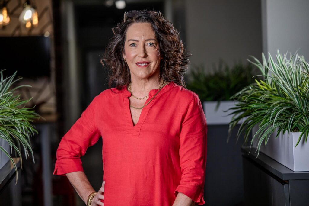 Elize Smith - The Brief | Namibia's Leading Business & Financial News Professional woman in a coral-red blouse standing with one hand on her hip in a modern office, plants in the background.