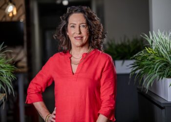 Professional woman in a coral-red blouse standing with one hand on her hip in a modern office, plants in the background.