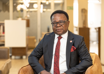Professional portrait of a man in a dark suit and red tie sitting in a beige chair in a modern office lounge.