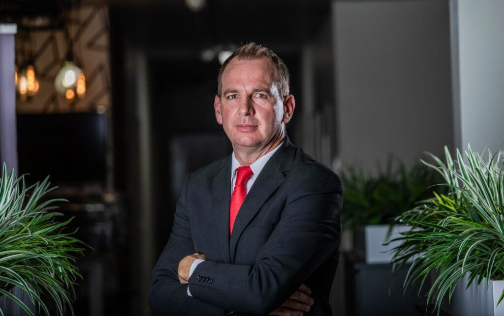 Hermanus Grobler - The Brief | Namibia's Leading Business & Financial News Portrait of a middle-aged man in a dark suit and red tie, arms crossed, standing between plants in a modern office setting.