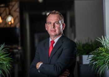 Portrait of a middle-aged man in a dark suit and red tie, arms crossed, standing between plants in a modern office setting.