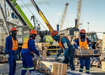 Group of workers in orange safety vests and hard hats handling cardboard boxes at a busy dock, with cranes and stacked pallets in the background.