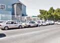 A row of white taxis parked along a street beside a blue industrial building, with a man standing near one car and trees in the background.