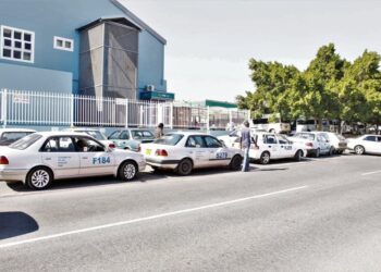A row of white taxis parked along a street beside a blue industrial building, with a man standing near one car and trees in the background.