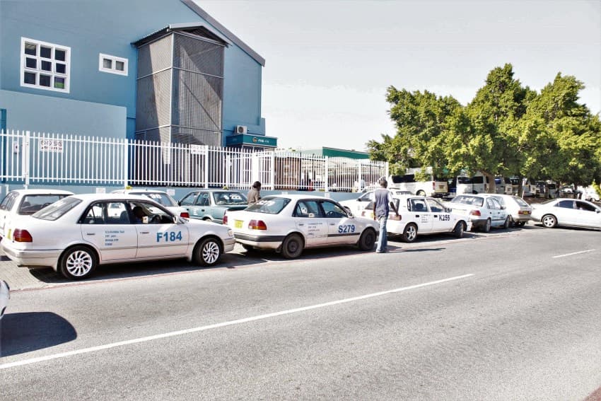 A row of white taxis parked along a street beside a blue industrial building, with a man standing near one car and trees in the background.