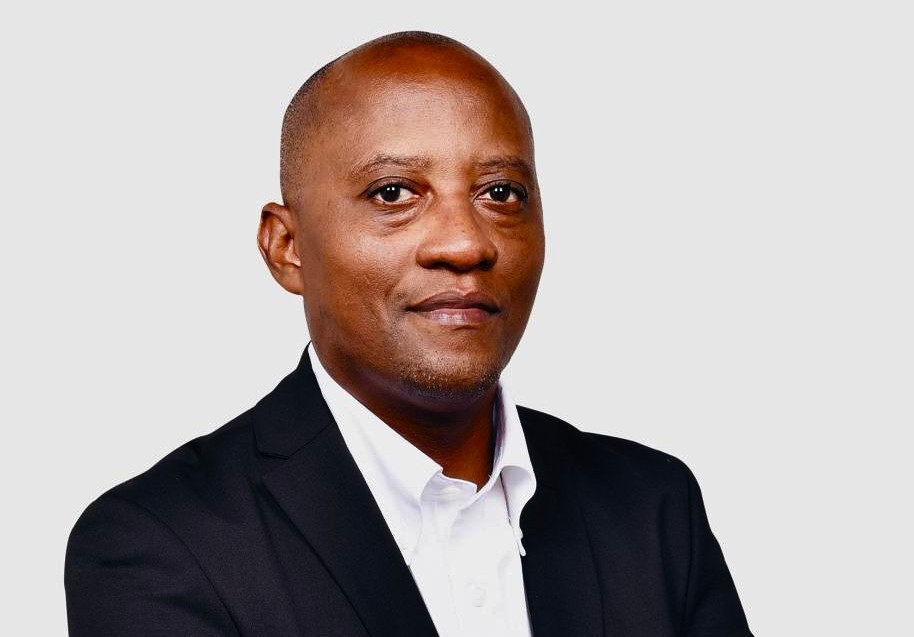 Professional headshot of a man in a black suit and white shirt against a light gray background, facing the camera.