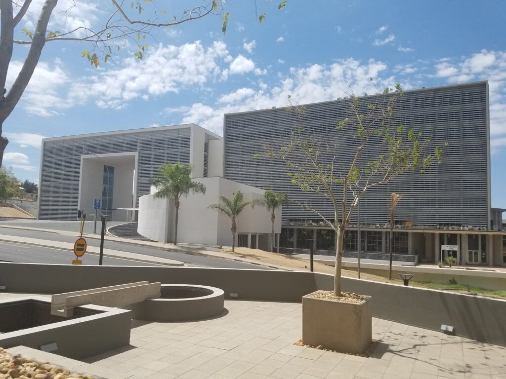 CITY OF WINDHOEK - The Brief | Namibia's Leading Business & Financial News White contemporary building with large metal louvers, palm trees, and a paved plaza under a blue sky with scattered clouds in the foreground.