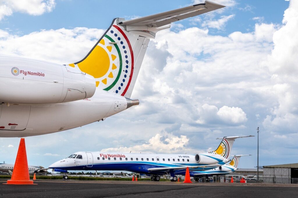 Airliner tail with colorful Fly Namibia logo on a tarmac beside orange traffic cones and another parked plane beneath a cloudy sky