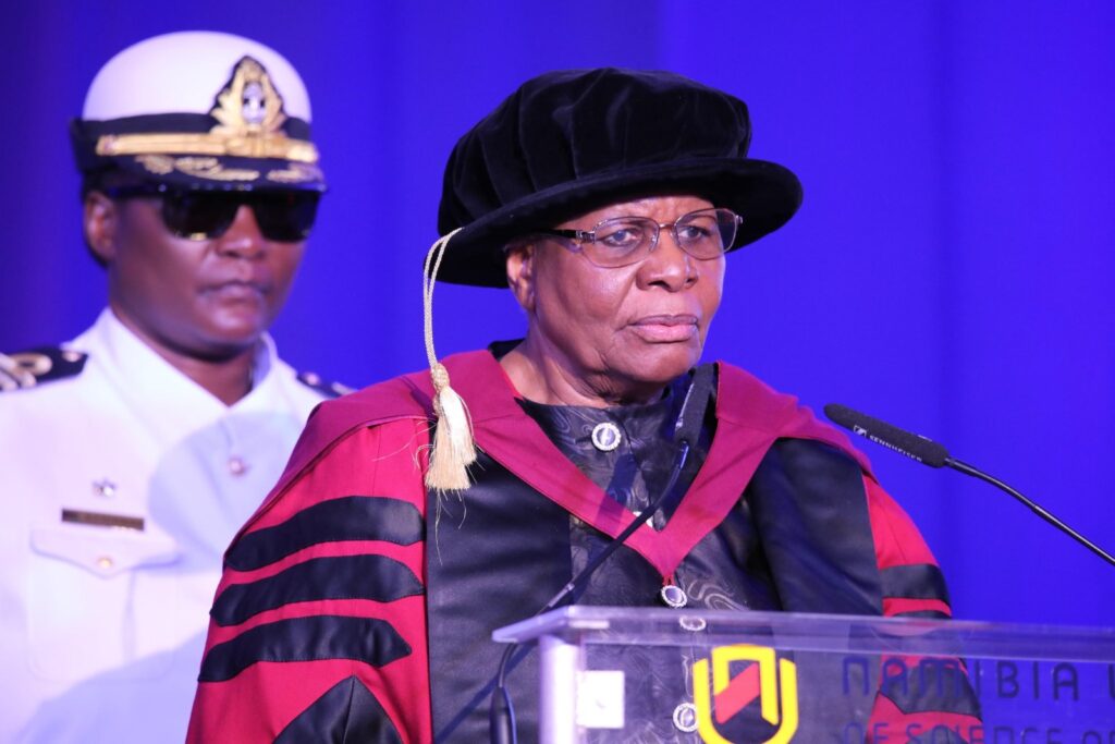 President Nust - The Brief | Namibia's Leading Business & Financial News Woman in academic regalia speaks at a podium during a formal ceremony, a uniformed man stands behind her against a blue backdrop.