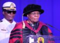 Woman in academic regalia speaks at a podium during a formal ceremony, a uniformed man stands behind her against a blue backdrop.