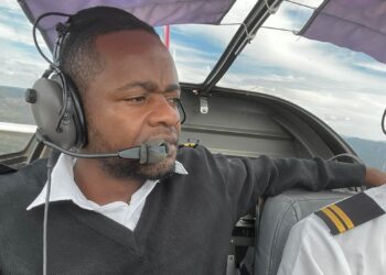 Male pilot wearing a headset and microphone in a small aircraft cockpit, looking focused at the horizon through the canopy.