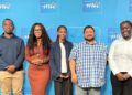 Group of five diverse individuals posing for a photo in front of a blue backdrop with the MTC logo at an event.