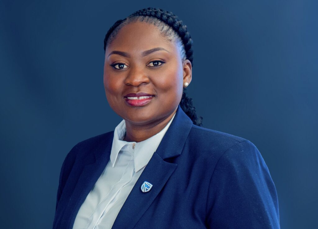 Hellen Amupolo - The Brief | Namibia's Leading Business & Financial News Professional headshot of a smiling woman in a navy blazer and white shirt against a blue background with a small lapel pin