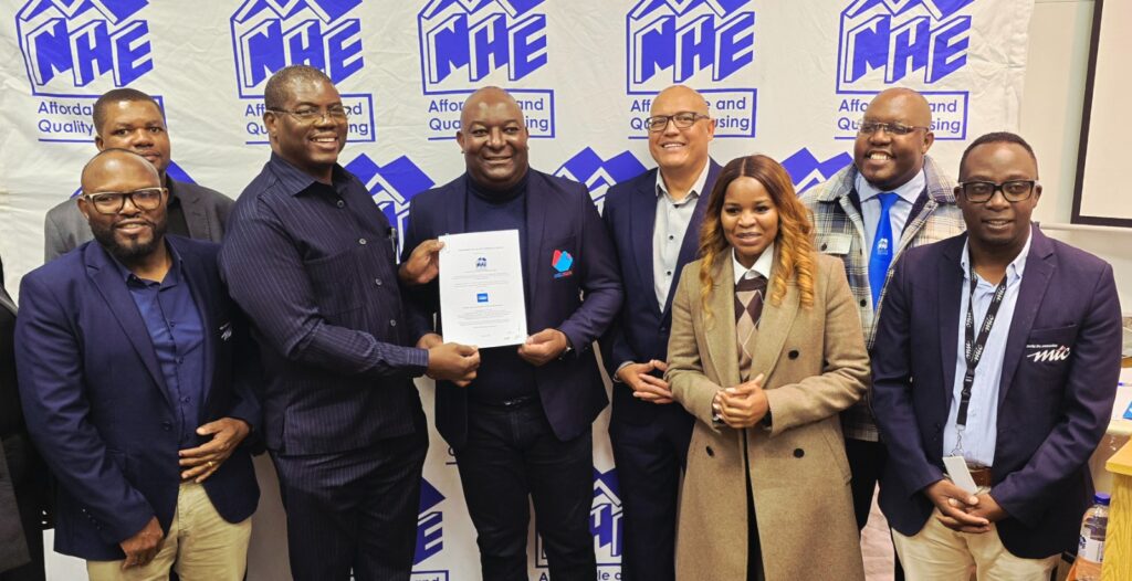 Eight professionals in business attire pose for a group photo, one man in the center holding a certificate in front of a blue and white NHE banner backdrop.