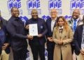 WhatsApp Image 2026-05-12 at 125751 PM - The Brief | Namibia's Leading Business & Financial News Eight professionals in business attire pose for a group photo, one man in the center holding a certificate in front of a blue and white NHE banner backdrop.