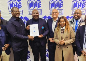 Eight professionals in business attire pose for a group photo, one man in the center holding a certificate in front of a blue and white NHE banner backdrop.
