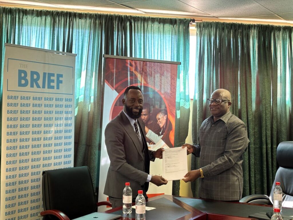 WhatsApp Image 2026-05-13 at 24514 PM - The Brief | Namibia's Leading Business & Financial News Two men in business attire exchange a document during a formal signing in a conference room, with banners behind them and water bottles on the table.