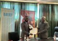 WhatsApp Image 2026-05-13 at 24514 PM - The Brief | Namibia's Leading Business & Financial News Two men in business attire exchange a document during a formal signing in a conference room, with banners behind them and water bottles on the table.
