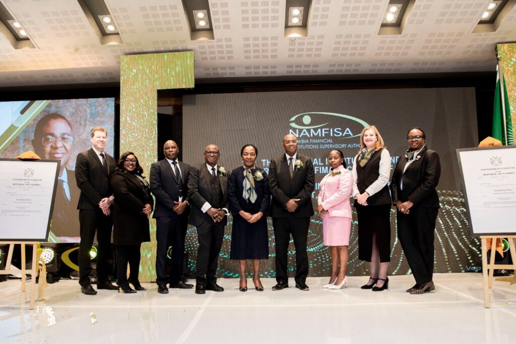 WhatsApp Image 2026-05-13 at 31216 PM - The Brief | Namibia's Leading Business & Financial News Nine formally dressed individuals pose on a stage during a ceremony, with a NAMFISA banner on a screen behind them and framed certificates on easels beside the group.